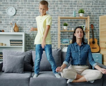 Happy kid boy is dancing on sofa while young mother is meditating in lotus position at home sitting on couch with closed eyes. Childhood and parenthood concept.