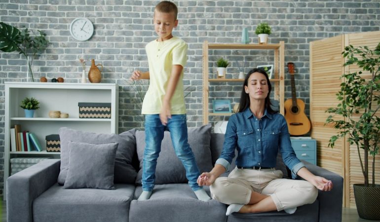Happy kid boy is dancing on sofa while young mother is meditating in lotus position at home sitting on couch with closed eyes. Childhood and parenthood concept.