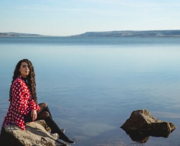 A woman in a red polka dot dress seated on rocks by a calm lake under a clear sky.