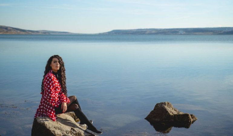 A woman in a red polka dot dress seated on rocks by a calm lake under a clear sky.